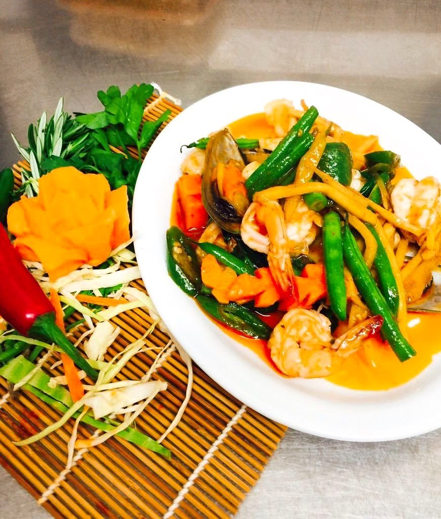 A stir fry of prawns, green beans, red chilli and vegetables in a white bowl, with carved carrot flowers and herbs on the side